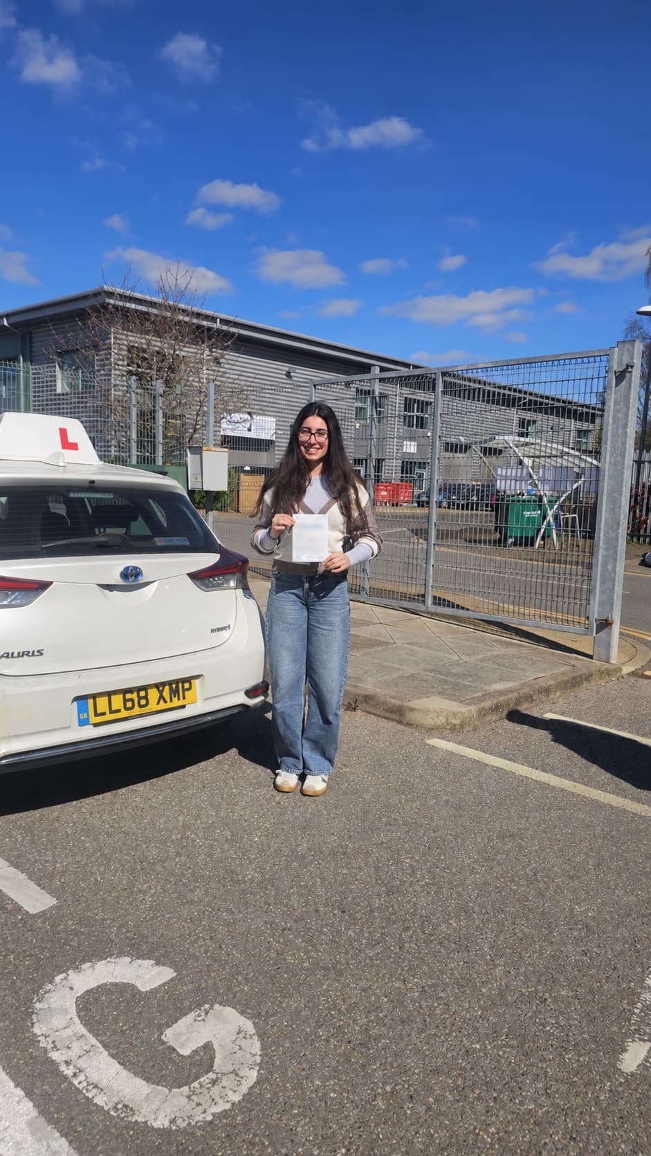 A pupil holding her pass certificate beside the Let's Go Driving car on a sunny day